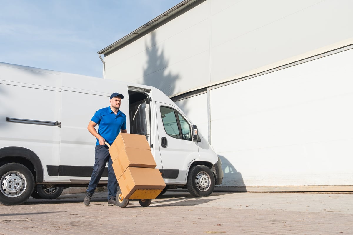 Delivery man in blue uniform transporting cardboard boxes on a hand truck outside a warehouse with a white van