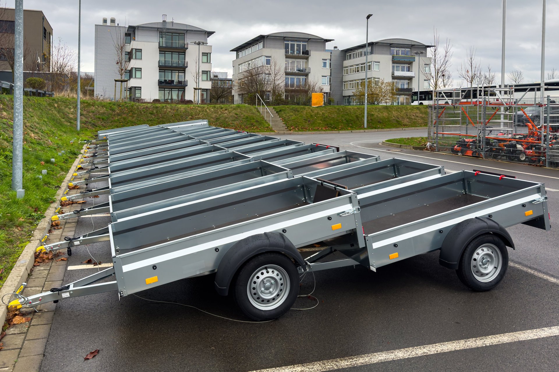 Several small trailers are lined up in a parking lot beside residential buildings. The weather appears cloudy. The trailers are ready for use or rental by individuals or businesses.