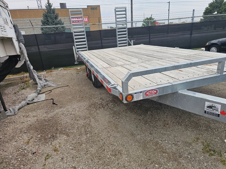 Empty aluminum flatbed trailer parked in an outdoor lot with industrial buildings and black fence visible in the background