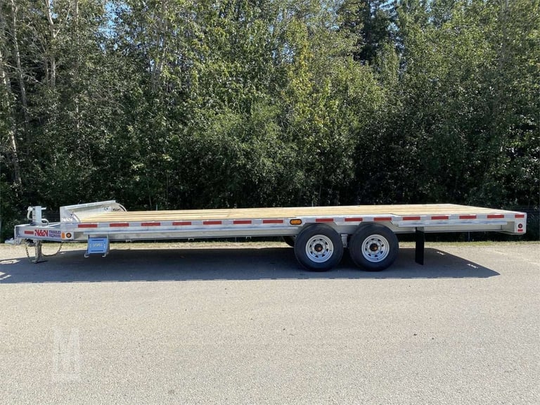 Empty aluminum flatbed trailer with dual axles parked on asphalt with evergreen trees in background