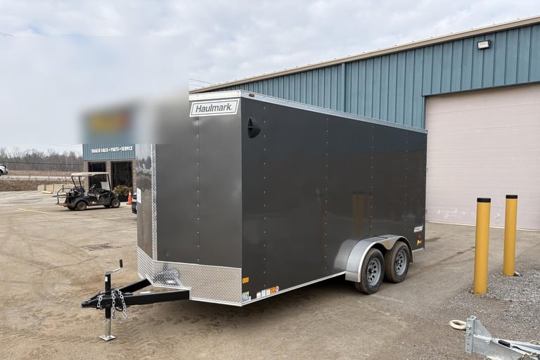 Black enclosed trailer with white trim and dual axles parked on gravel lot with warehouse building in background